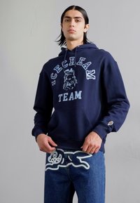 Navy blue hooded sweatshirt with "ICE CREAM TEAM" print, featuring contrasting light blue graphics and front pocket. Worn with denim jeans.