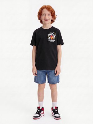 Dreng med rødt hår iført sort T-shirt med grafisk tryk, blå denimshorts, hvide sokker og sorte, hvide og røde sneakers, står og smiler.