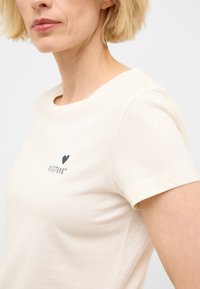 Camiseta de color crema hecha de algodón suave, con un escote redondeado y mangas cortas. Incluye un gráfico de un corazón y el logo "MUSTANG".