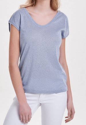 Femme portant un haut à manches courtes bleu clair avec un col en V et un pantalon blanc, debout devant un fond blanc uni.