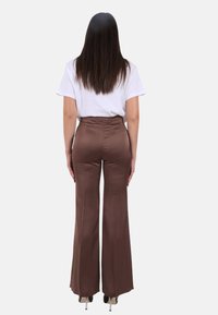 Pantaloni marroni a vita alta e gamba svasata con una texture liscia, abbinati a una t-shirt bianca semplice. Modello di spalle, che mostra scarpe con tacchi trasparenti.