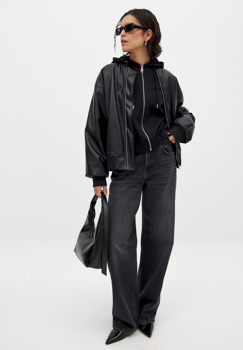 Black faux leather jacket over a black zip-up hoodie, paired with dark jeans and pointed black heels. Accessories include a black bag.