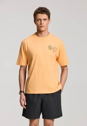 Jonge man draagt een perzikkleurig T-shirt met korte mouwen en palmprint en zwarte korte broek, staand tegen een eenvoudige lichte achtergrond.