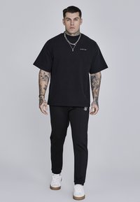 Črna oversized majica in črne tapered hlače z logotipnimi poudarki, v kombinaciji z belimi športnimi copati, z gladko bombažno teksturo in sproščenim fitom.