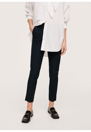 Persoon draagt zwarte slim-fit broek, wit oversized overhemd en zwarte loafers met gouden gesp, staande met één hand in de zak.