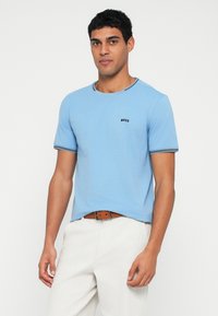 Jasnoniebieska koszulka z krótkim rękawem i okrągłym dekoltem, z czarnymi akcentami na kołnierzu i rękawach oraz małym czarnym logo.