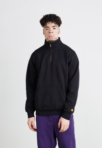 Sort fleece trøje med kvart-zip krave, frontlomme og gul logoaccent på ærmet, parret med lilla bukser.