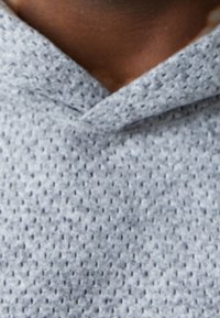 Ljusgrå huvtröja i texturerad tyg med V-ringning, som har ett mjukt och andningsbart stickat mönster. Ingen synlig hårdvara eller detaljer.