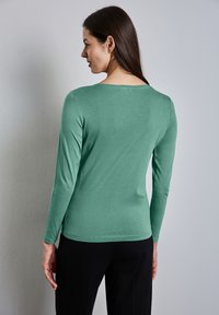 Pitkähihaisessa pehmeästä vihreästä kankaasta valmistetussa paidassa on joustava muotoilu, pyöreä pääntie ja sileä pinta, yhdistettynä mustiin housuihin.