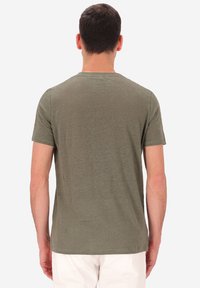 Uomo in piedi di spalle, che indossa una maglietta verde oliva a maniche corte e pantaloni bianchi, su uno sfondo bianco semplice.