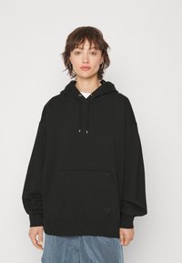 Sudadera negra de corte holgado hecha de tejido suave, con un bolsillo frontal, capucha con cordón y detalles en dorado. Presenta un logo en el dobladillo.