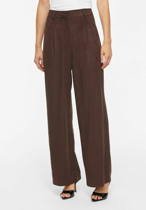Femme portant un pantalon marron taille haute à jambes larges associé à des talons ouverts noirs sur fond blanc.