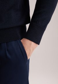 Granatowy sweter z dzianiny z prążkowanymi mankietami, w połączeniu z gładkimi granatowymi spodniami. Ręka oparta w kieszeni, podkreślająca teksturę i kontrast kolorów.