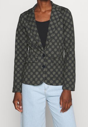 Person iført en mønstret sort og grå blazer over en sort top, kombineret med lyseblå jeans, stående mod en ensfarvet baggrund.