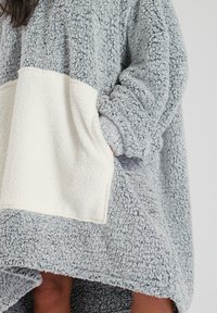 Sudadera de felpa gris de gran tamaño con un bolsillo crema, exterior texturizado, mangas amplias y un diseño de dobladillo asimétrico.