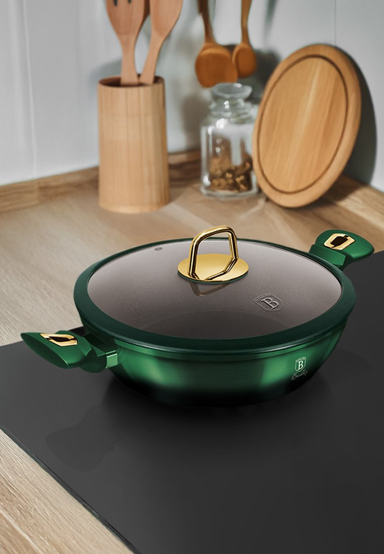 Pot en cuisson vert avec couvercle en verre, poignée dorée et accents latéraux. Fini lisse. Posé sur une surface noire avec des ustensiles de cuisine en bois en arrière-plan.