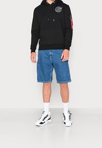 Sudadera negra con un bolsillo frontal y un logo circular, combinada con pantalones cortos de mezclilla azul, calcetas blancas y zapatillas de deporte en blanco y negro.