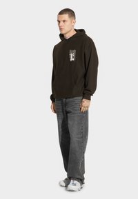 Ciemnobrązowa bluza z kapturem z kieszenią z przodu i grafiką na lewej piersi, w połączeniu z szarymi, luźnymi dżinsami i białymi trampkami.