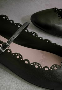 Ballerine in pelle nera con bordi scallop, ritagli decorativi e cinturino alla caviglia. Fodera interna rosa. Texture liscia.