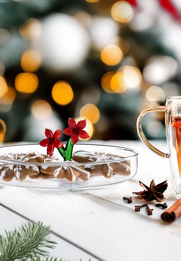 Durchsichtiger Glasplatte mit braunen Lebkuchenkeksen, weißem Zuckerguss und einer roten Blütendekoration; ein Glasbecher mit orangefarbener Flüssigkeit neben Zimt und Sternanis.