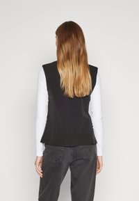 Svart ärmlös väst med en tight passform, tillverkad av slät tyg, bärs över en vit långärmad topp och kombineras med mörkgrå jeans.