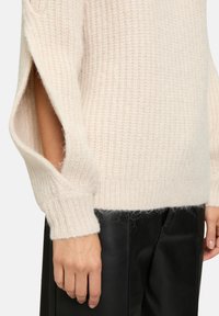 Maglione a maglia color beige chiaro con polsini a coste, dotato di aperture laterali. Il tessuto morbido e testurizzato contrasta con i pantaloni in pelle nera liscia.