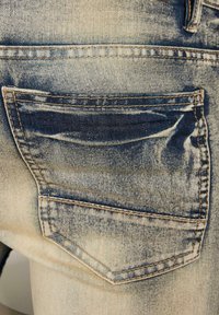 Denimjeans med en falmet blå vask, som har en baklomme, kontrastfarget søm, og en teksturert overflate med diskret slitasje.