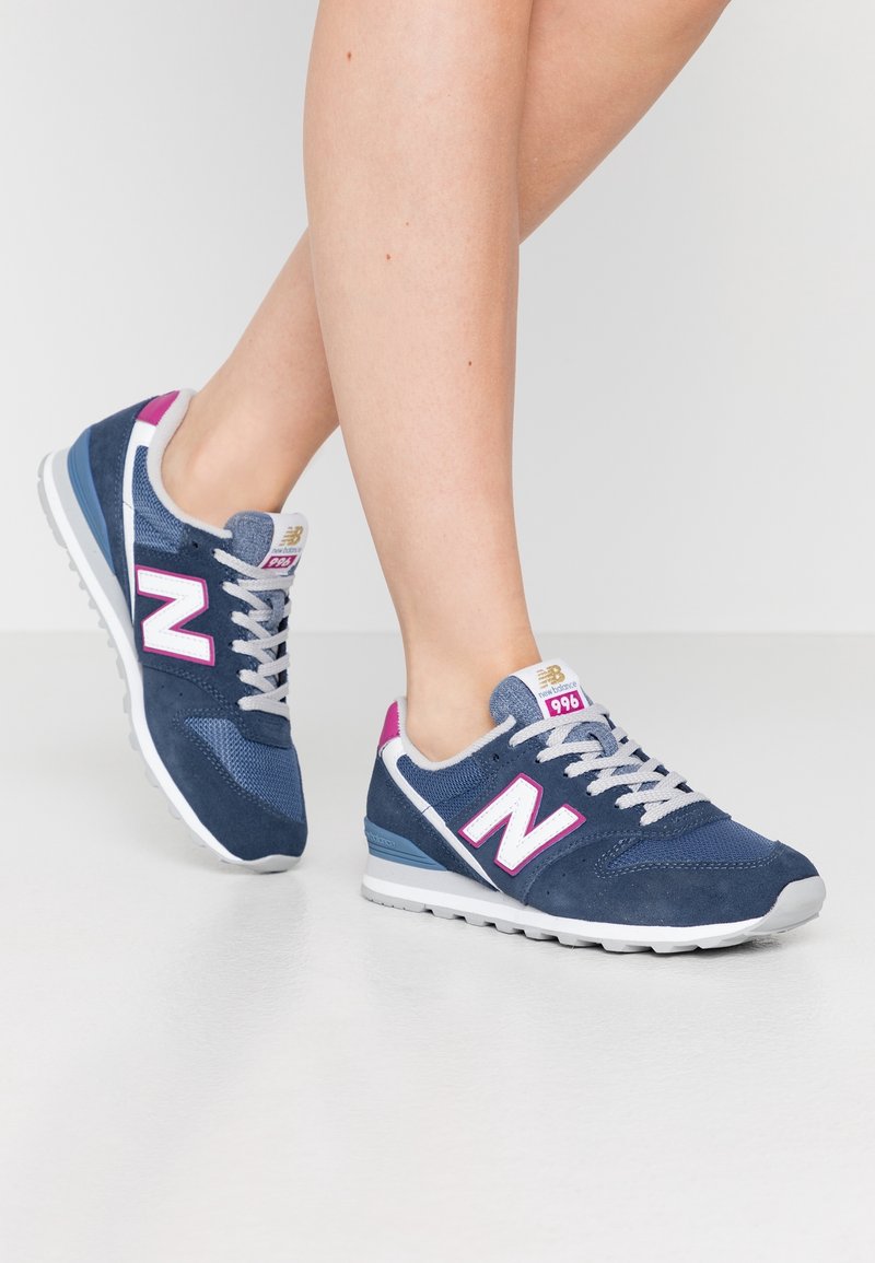 Baskets en daim bleu et en mesh avec une semelle blanche, arborant un grand logo "N" rose, des lacets gris et un accent rose sur le talon.
