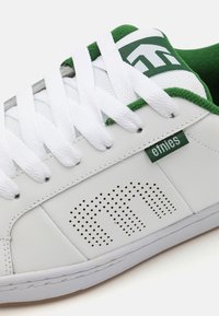Tenis brancos com detalhes em verde, apresentando pormenores perfurados, atacadores texturizados brancos e um colarinho almofadado. Etiqueta da marca na lateral.