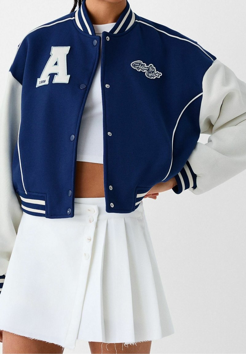 Marinblå och vit varsity-jacka med ett "A"-logotyp, knappar och randiga ribbade ärmslut. I kombination med en vit plisserad kjol.
