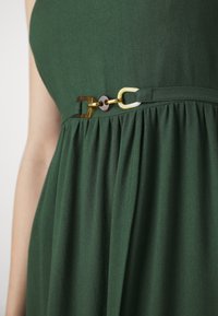 Vestido verde escuro com textura franzida e um cinto decorativo com detalhes em dourado, castanho e marfim.