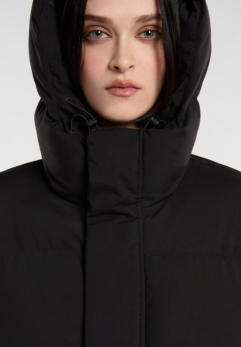Chaqueta de plumas negra con cuello alto y capucha ajustable, con textura acolchada y detalles cosidos para mayor aislamiento.