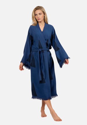 Frau trägt einen blauen, knielangen Kimono-ähnlichen Bademantel mit schwarzen Dreiecksmustern, weiten Ärmeln, fransigen Kanten und einem gebundenen Taillengürtel.