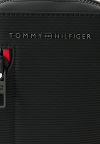 Bolsa de nailon negro texturizado con la marca Tommy Hilfiger. Cuenta con cierre de cremallera y un acento interior de color contrastante.