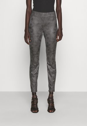 Vrouw die strakke grijze, gestructureerde broek met een hoge taille en zwarte sandalen met bandjes en hoge hakken draagt, staand tegen een effen achtergrond.