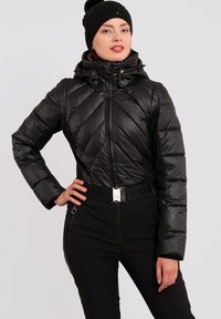 Zwarte pufferjack met een slank ontwerp, ruitpatroon, hoge kraag en ritssluiting. Gecombineerd met zwarte broek en een gebreide muts met textuur.