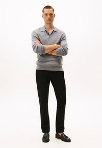 Mees seisab käed rüseluses, kandes halli pika varrukaga polo, musti pükse ja musti tuttnahkseid mokasiine, taustaks on lihtne valge sein.