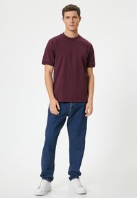 T-shirt a maniche corte color maroon, collo a giro, vestibilità rilassata, abbinato a jeans in denim blu e scarpe da ginnastica bianche, in piedi su uno sfondo neutro.