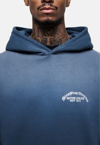 Felpa blu realizzata in un tessuto morbido, con tasca frontale, cappuccio con coulisse e logo ricamato bianco sul petto.