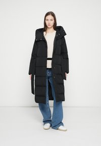 Mladá žena oblečená v dlouhém černém péřovém kabátě, béžovém svetru, modrých širokých džínách a bílých teniskách stojící před jednobarevným bílým pozadím.