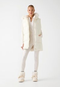 Veste blanche matelassée avec capuche en fausse fourrure, portée sur un body léger à motif et associée à des bottes crème. Texture lisse et fermeture éclair.