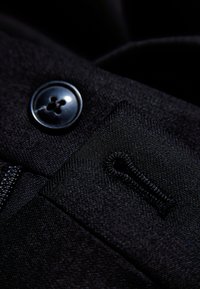 Zwarte stof close-up met een ronde knop met vier gaten, gestikt detail en een kleine stoffen lus. Gladde textuur, consistente kleur.