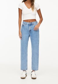 Vaqueros azul claro de talle alto con piernas rectas, que incluyen trabillas para cinturón y cinco bolsillos, combinados con un top corto blanco de manga corta y zapatillas deportivas.