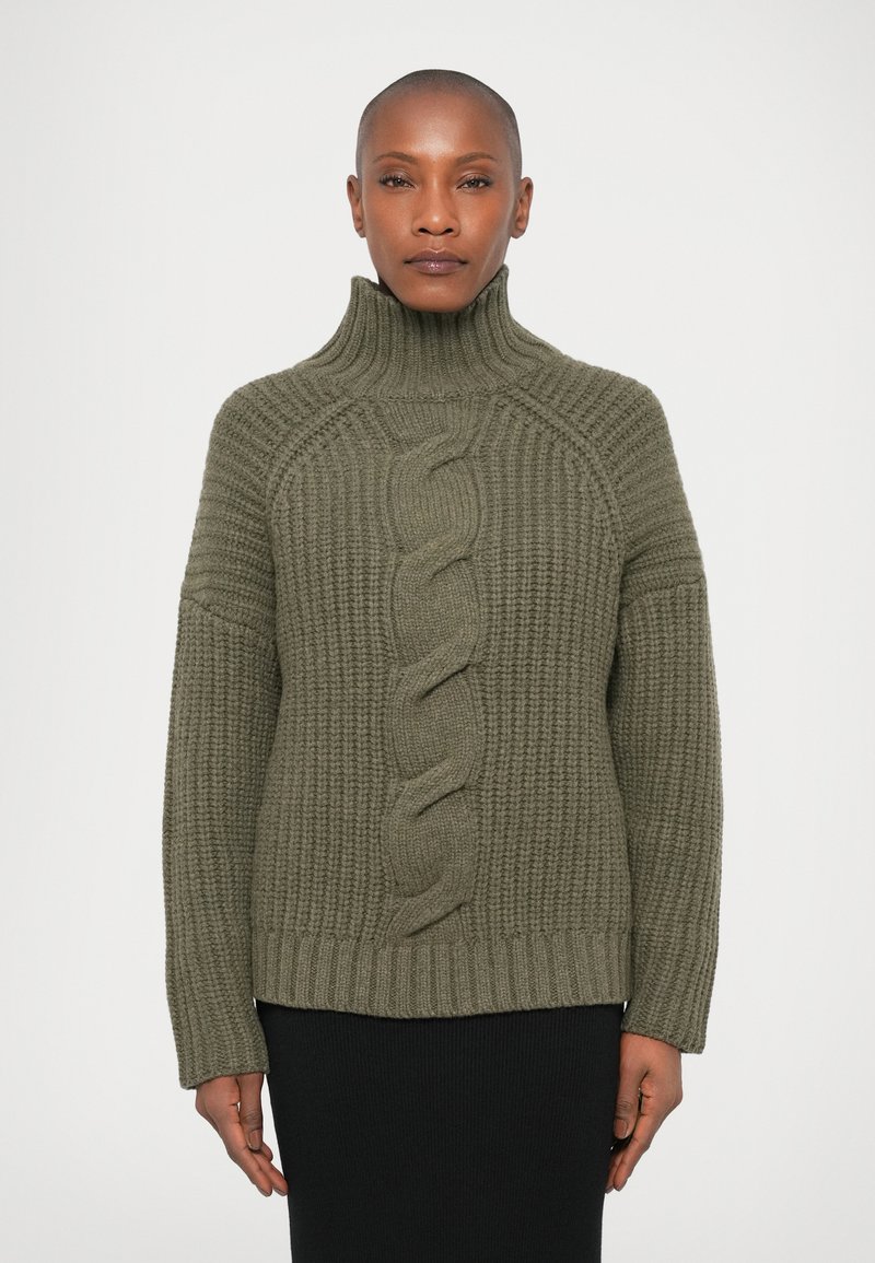 Mulher vestindo um suéter de tricot grosso em verde oliva com um padrão trançado central, combinado com uma saia preta, em pé contra um fundo branco.