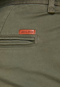 Pantaloni di cotone verde oliva con un'etichetta di pelle marrone visibile con la scritta "JACK & JONES" e un dettaglio discreto di tasca laterale.