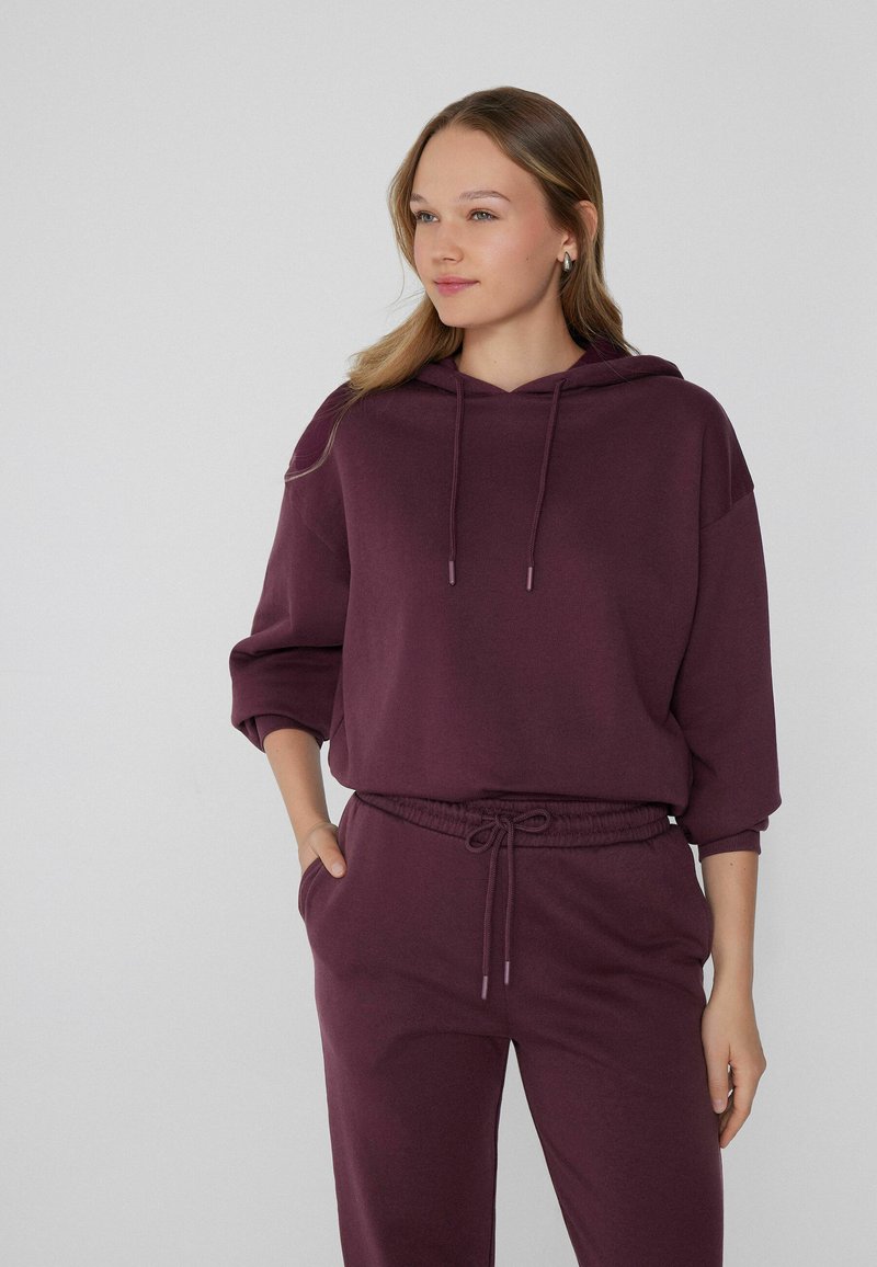 Bordowy hoodie z sznurkiem, obniżonymi ramionami i luźnym fasonem, w połączeniu z pasującymi dresami z elastycznym paskiem i kieszeniami.