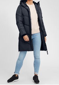 Černý, podšitý dlouhý kabát s kapucí, který má zipové uzavírání a elastické manžety. Nalezeno přes lehký svetr a modré džíny. Černé tenisky.