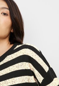 Gestrickter Pullover mit schwarzen und goldenen Streifen, der über einen V-Ausschnitt und ein strukturiertes Muster verfügt. Fokus auf horizontalen Streifen und Nähdetails.