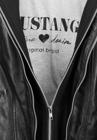 Schwarze Lederjacke mit silbernem Reißverschluss, darüber ein graues Shirt mit schwarzem Schriftzug und Herzdesign, auf dem "MUSTANG pure denim" steht.