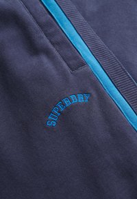 Close-up van donkerblauwe stof met blauwe bies en "SUPERDRY" geborduurd in het blauw boven een opening van de zak.